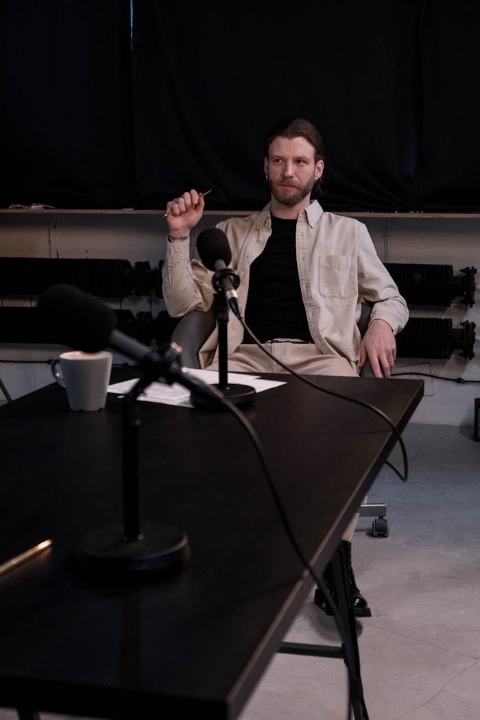 Man in a studio sitting at a table for a podcast interview with microphones.